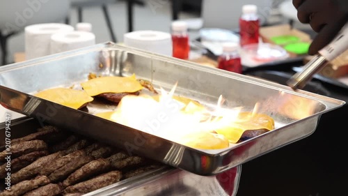 Professional chef uses cooking torch to melt yellow cheese on grilled burger patties in commercial stainless steel catering tray for outdoor party buffet fresh food preparation service.