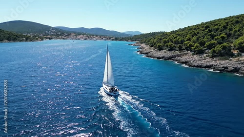 White sailboat cruising blue sea near forested island coastline.