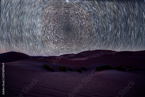 View of a lone person standing on a sand dune under a dramatic circular star trails in the night sky.