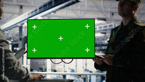 Fotografía Soldier and female technician use a green screen computer in a server hub to manage systems with a mockup workstation