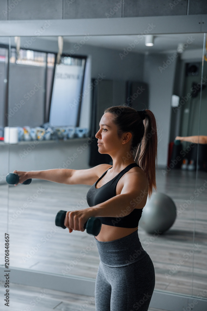 Fototapeta premium Young woman training with dumbbells during energetic indoor fitness exercise routine. Pretty woman lifting hand weights with athletic balanced posture inside studio. Strength wellness concept.