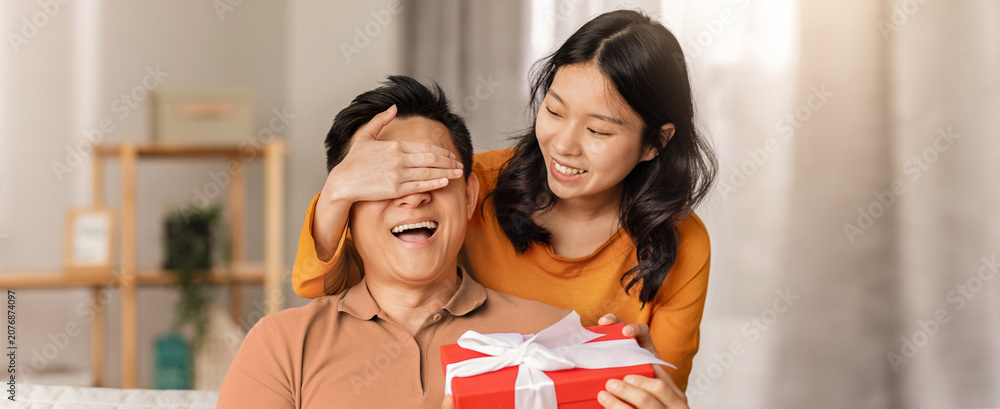 Obraz premium Joyful Emotion. Happy Young Asian Woman Surprising Man With Gift Box While Covering His Eyes, Celebrating A Special Occasion Together At Cozy Home Setting. Family Concept.