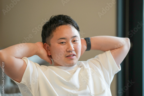 Man rests hands behind his head. He wears a simple white tee. Soft lighting highlights his calm expression. Neutral background keeps focus on his relaxed pose. Ideal for wellness, relaxation