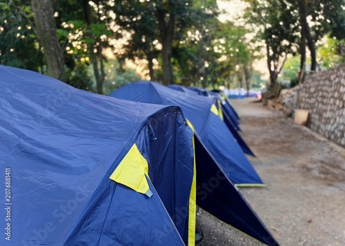 Row of blue camping tents set up in a row at an outdoor campsite surrounded by trees.