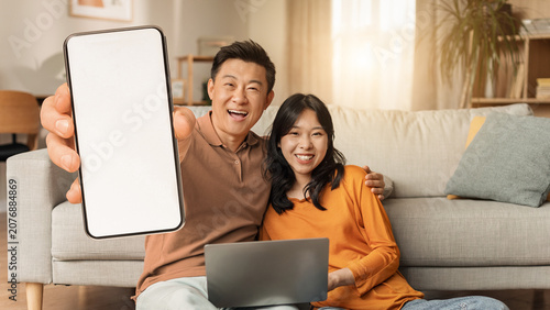 Tableau sur toile Happy Asian Couple, Male And Female In Their 30s, Smiling Together While Holding Smartphone With Blank Screen, Sitting On Couch In Cozy Living Room, Enjoying Lifestyle And Technology Concept