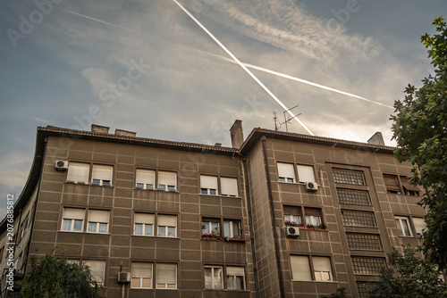 Residential apartment block in Belgrade, Serbia, showing a concrete facade, window shutters, air conditioners, balconies, and evening sky with aircraft contrails above the urban housing complex.