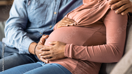 Fotografie Happy Pregnant Black Woman Touching Her Belly With Partner, Sitting Together On Couch At Home, Sharing Joyous Moment Of Expectation And Love In Cozy Setting