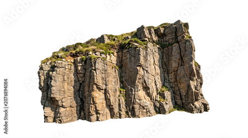 Low angle view of a massive, rugged rocky cliff face with sparse green vegetation on top against a white sky.