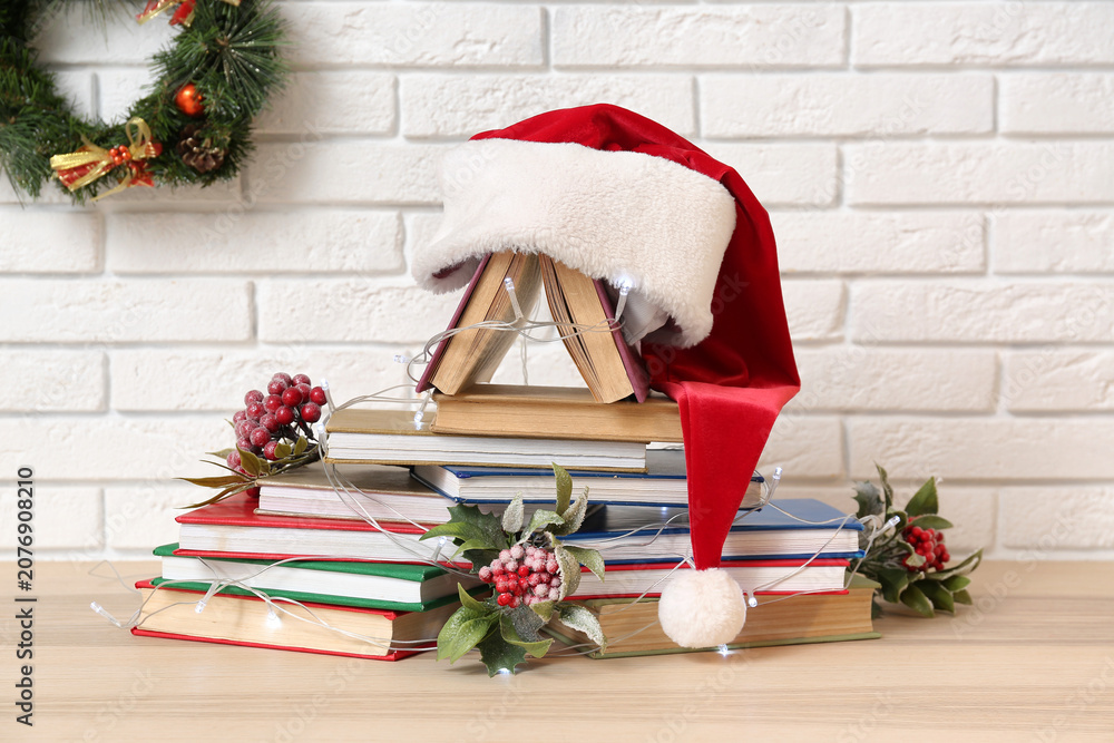 Fototapeta premium Christmas tree made of books, berries, glowing lights and Santa hat on table near white brick wall
