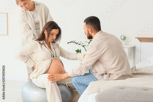 Young pregnant woman sitting on fitness ball with her husband and doula in be...