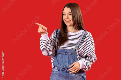 Quadro su tela Laughing young woman pointing at something on red background