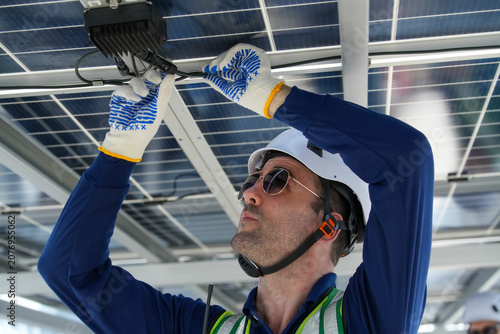 Engineer maintenance hvac cooling tower solar panel technician wiring electrician works with calm focus under array, adjusting cable, wearing helmet, glove, safety vest, outdoor daylight energy