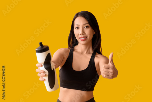 Beautiful young Asian woman with bottle of water showing thumb-up gesture on ...