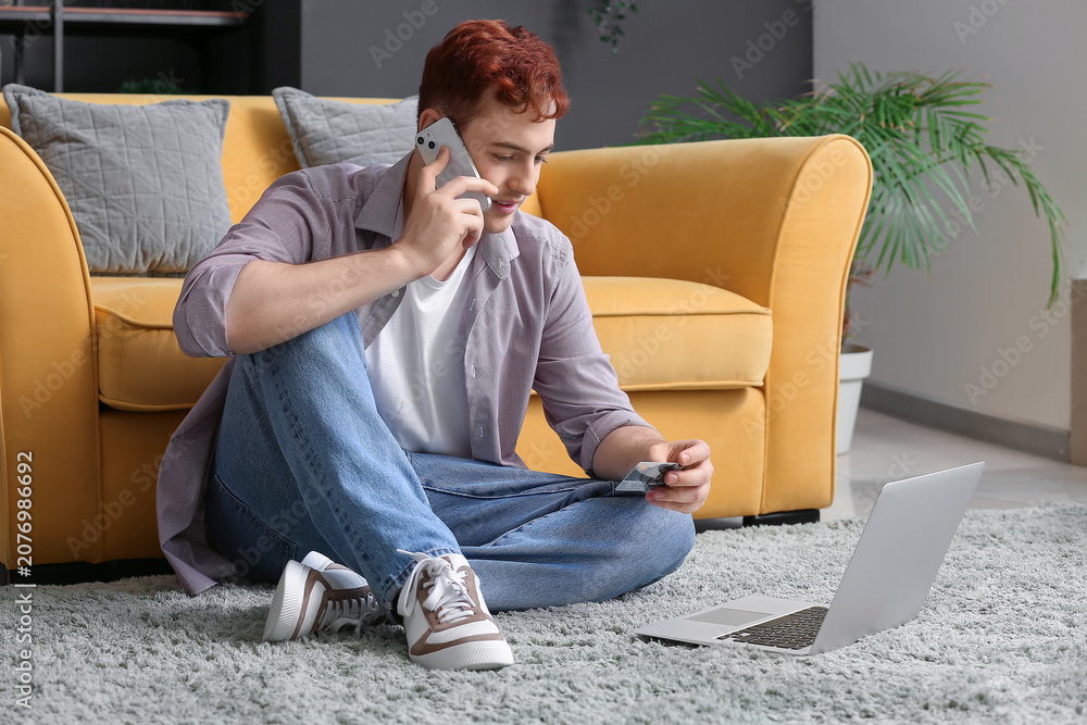 Fototapeta premium Handsome young man with credit card, mobile phone and modern laptop sitting on floor near sofa while shopping online