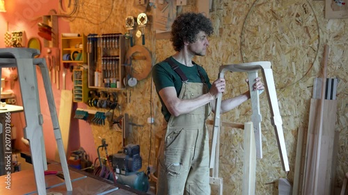 Private production of wood products, cheerful focused man carpenter examines semi-finished chair, craftsman holding stool and precisely checking quality of wooden elements, crafter proud of new item.