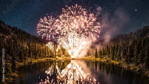 Fireworks explode over serene lake on USA Independence Day evening surrounded by trees and mountains.