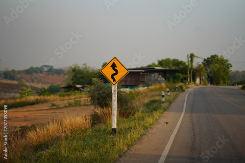 Yellow winding road warning sign beside a rural asphalt highway with countryside landscape and dry grass. Transportation safety concept for driving, navigation, travel.
