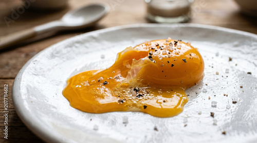 Close up of soft boiled runny egg yolk broken open on white ceramic plate seasoned with sea salt crystals and cracked black pepper rustic wood table