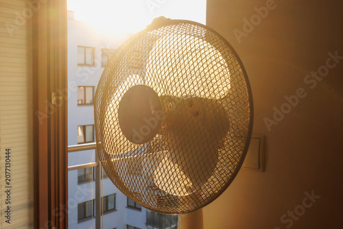 Slika na platnu An electric fan standing inside a room at sunrise on a hot summer morning, circulating cool air to improve ventilation during an extreme heatwave