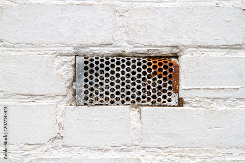 Metal ventilation grate embedded in a white painted brick wall. Industrial air vent cover with rust on a masonry surface. Building exterior maintenance and infrastructure concept.