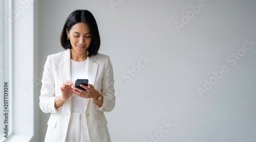 Smiling businesswoman in white blazer using smartphone against plain gray wall with large copy space, professional corporate portrait for business marketing