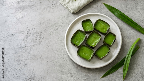 Top view of traditional green pandan dessert in banana leaf cups on a white plate. Grey stone background with copy space. Perfect for Asian culinary, recipe blogs, and food photography concepts.