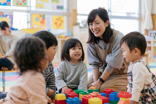 カラフルなブロックの周りで床に座る幼児たちと笑顔で話す日本人保育士