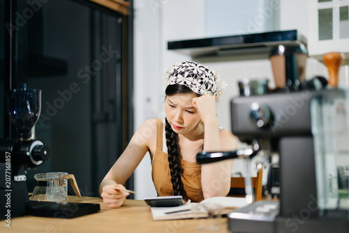 A worried female barista looks at a credit card while using a calculator at the counter. Concept of small business debt, financial stress