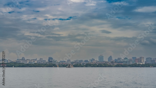 Beautiful calm urban lake. The ship is sailing. There is green vegetation on the shore, multi-storey city buildings. Skyscrapers against the sky and clouds. China. Hangzhou. West Lake Xi Hu   