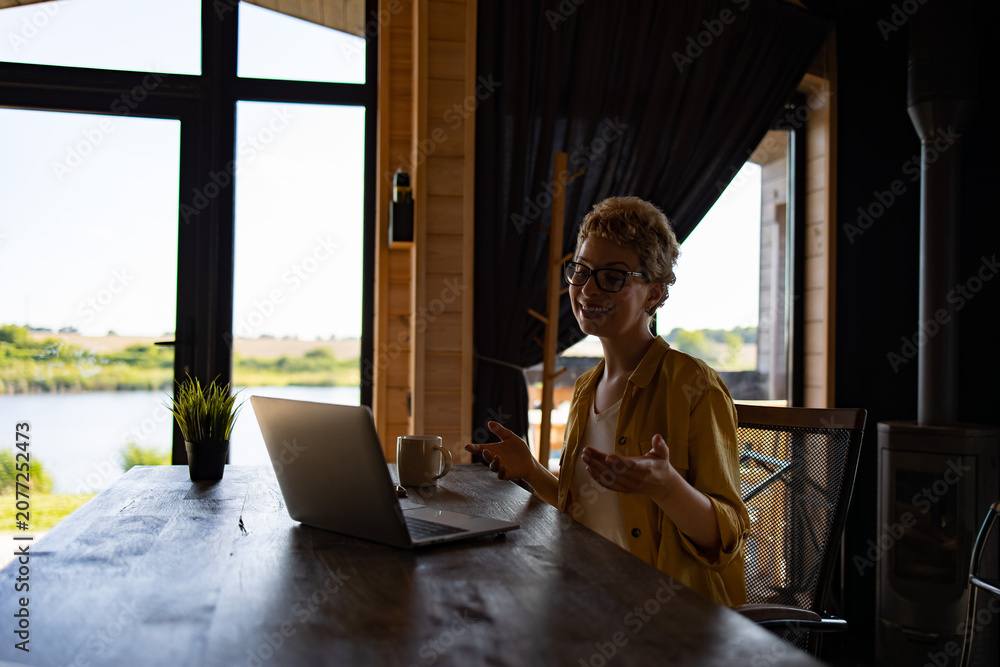 Fototapeta premium A Caucasian woman with glasses smiles while gesturing during a video call on her laptop at a wooden table with a scenic lake view