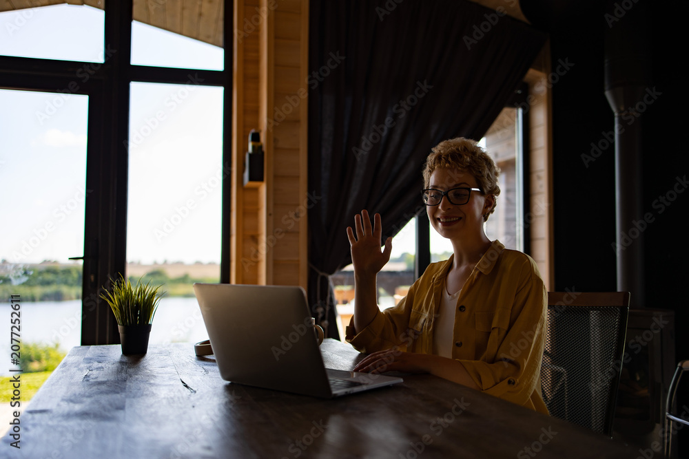 Fototapeta premium A smiling Caucasian woman with glasses waves hello while on a video call on her laptop, seated at a wooden table with a scenic view outside