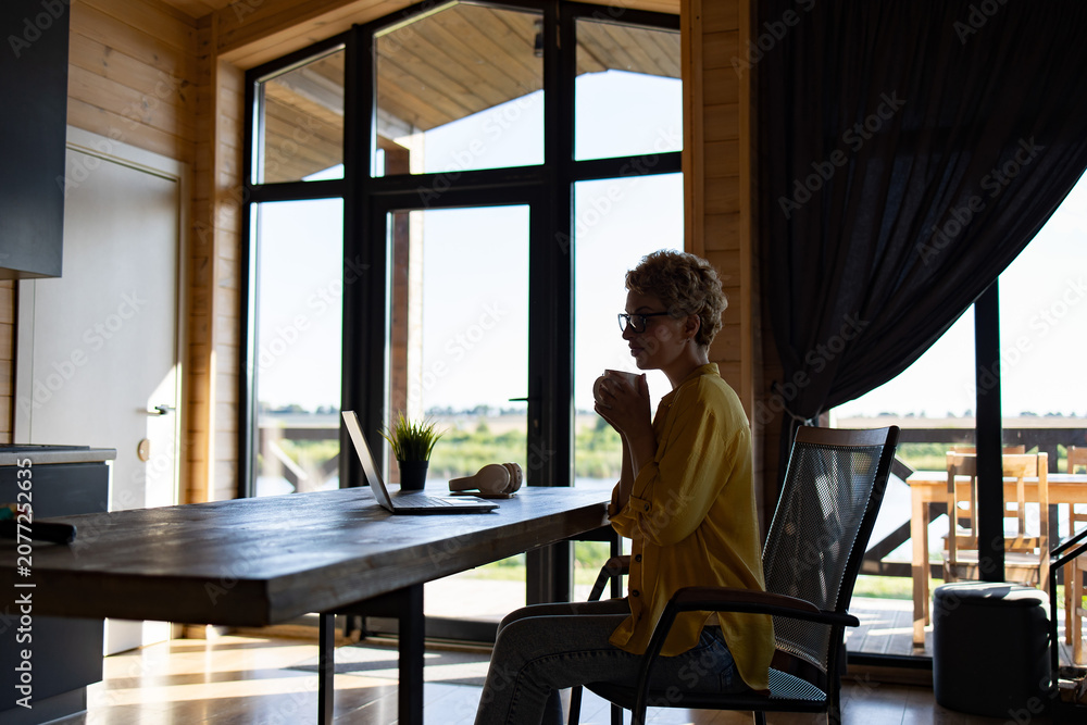 Fototapeta premium A Caucasian woman enjoys a hot beverage while working on her laptop in a modern wooden cabin with large windows overlooking a scenic landscape