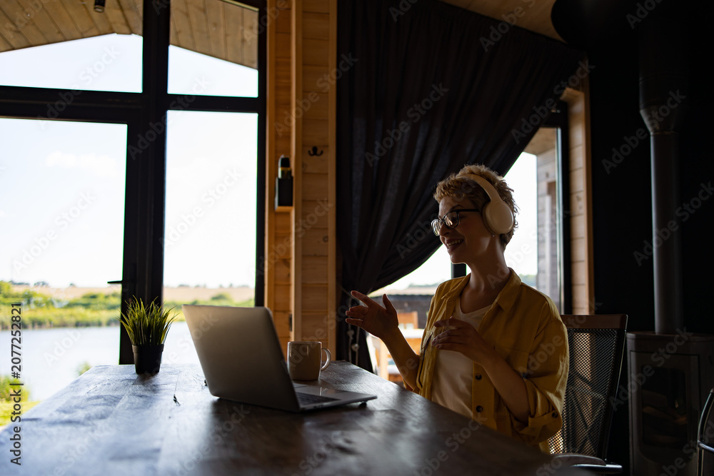 Fototapeta premium A Caucasian woman wearing headphones and glasses smiles while working on her laptop at a wooden table with a scenic view outside the window