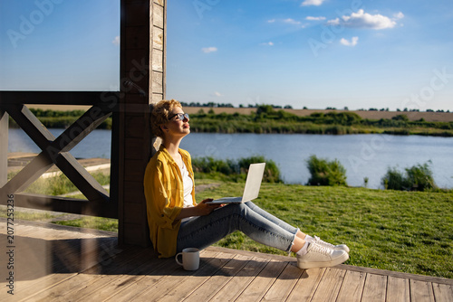 A Caucasian woman enjoys a sunny day while working on her laptop on a wooden porch overlooking a tranquil lake and green landscape