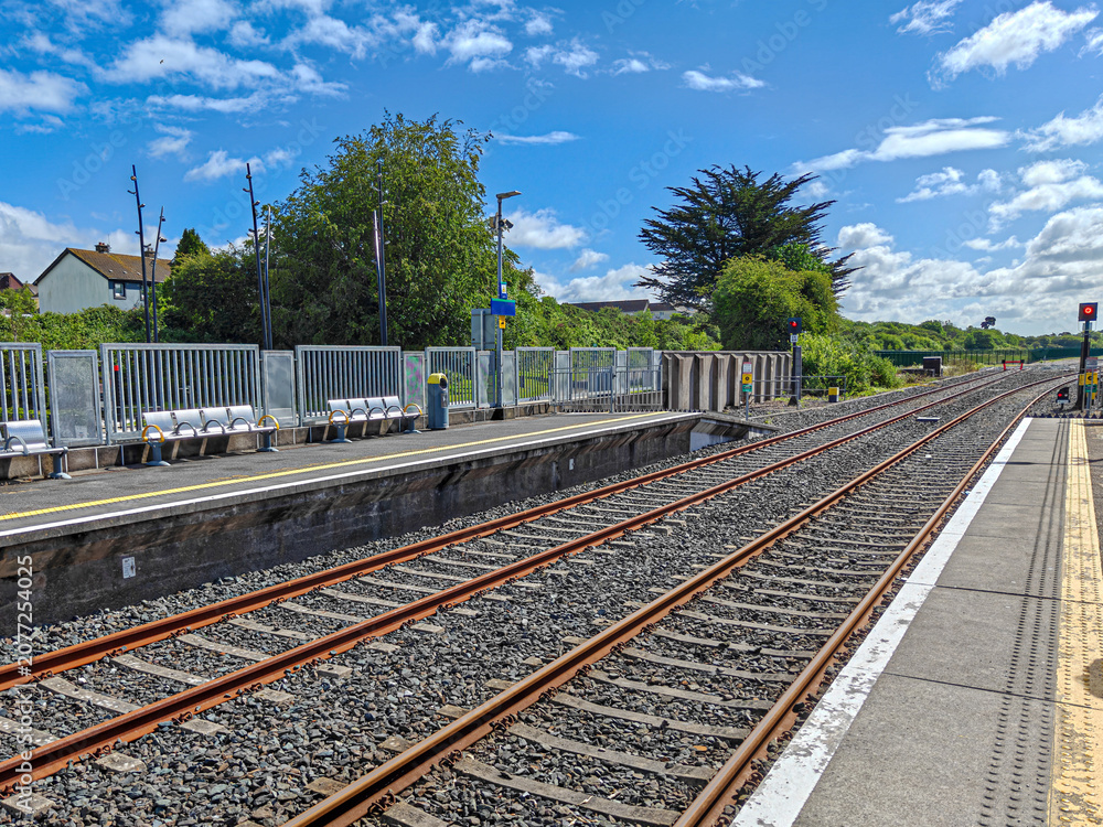 Fototapeta premium Train station with dual railway tracks extending into the distance. The tracks are bordered by gravel, and the platform has safety markings. Metal railings line the platform edge, with a few benches f