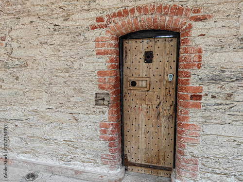 Narrow wooden door with metal studs set in an arched red brick and stone wall. The wood is weathered, and there's a small inspection hatch and a metal lock near the top. 