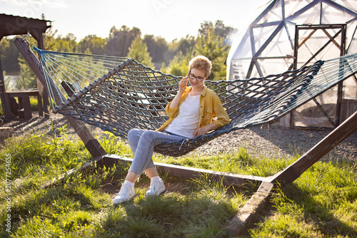 A young Caucasian woman with short blonde hair and glasses is talking on her phone while relaxing in a hammock on a sunny day in a grassy yard with a geodesic dome structure in the background