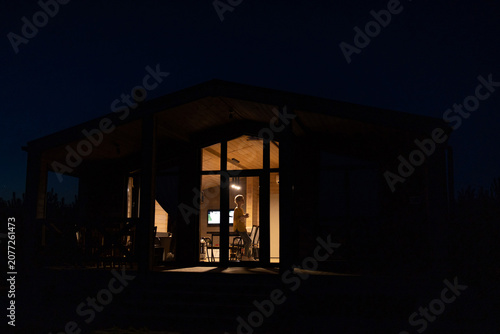 A solitary caucasian woman stands inside a modern cabin illuminated by warm interior lights against the dark night sky