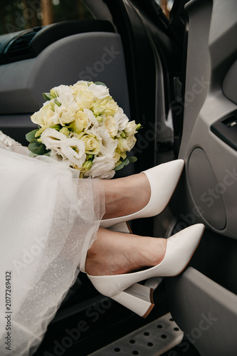 The slender legs of a young bride wearing white high-heeled shoes look out of the open car door next to a bouquet of wedding flowers