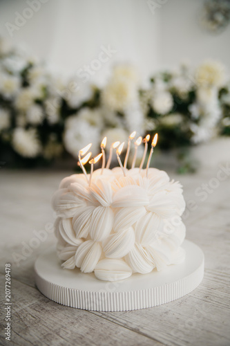 A birthday cake with burning candles inserted into its top stands against a background of flowers in a photo studio