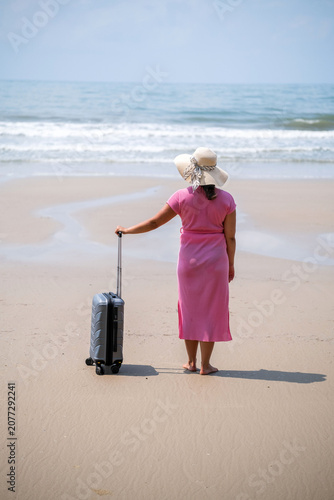 Young woman in pink dress and wide straw hat standing with a grey suitcase at the edge of the sea with waves lapping nearby on a sandy beach. Shoreline travel concept