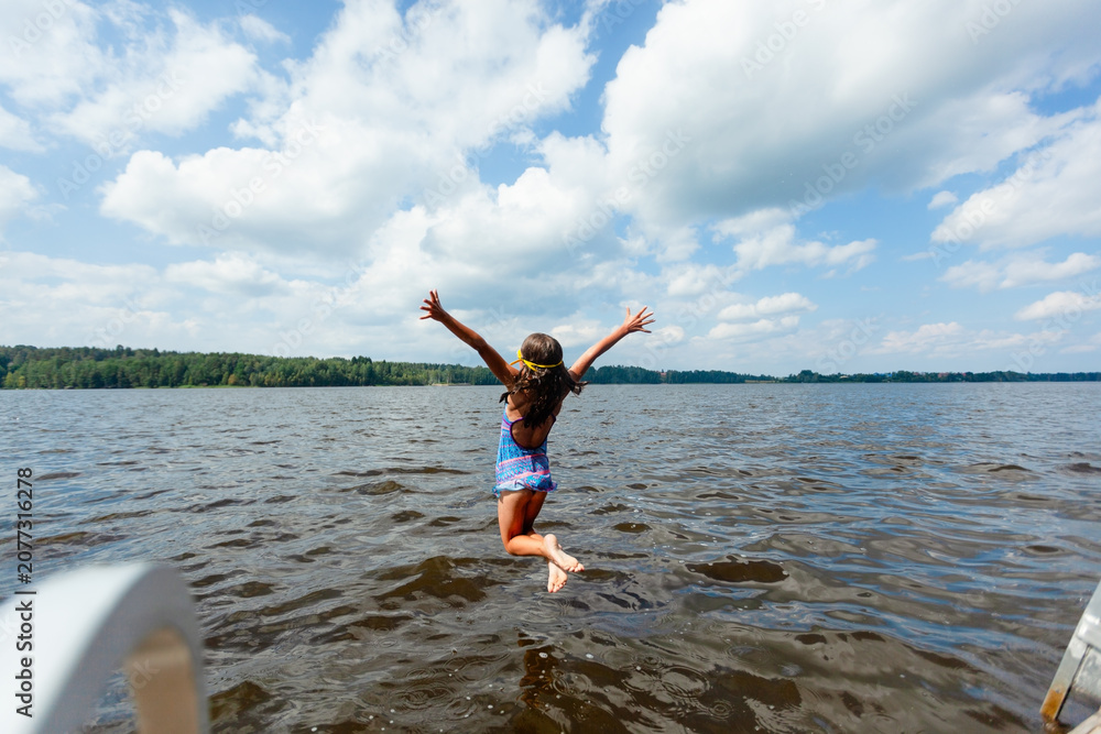 Obraz premium Girl jumps into a lake from a wooden dock on a sunny summer day. Child in swimsuit full of energy and excitement splashing into the water. Kid enjoys thrilling outdoor recreation and refreshing swim