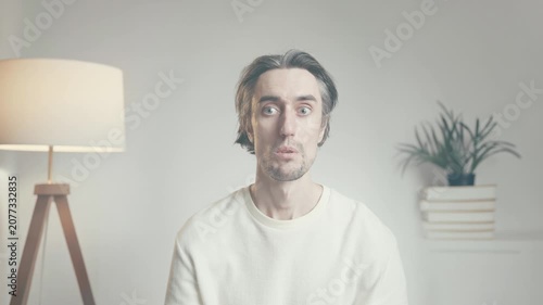 Young man with long hair showing surprise, confusion, and contemplation indoors in bright minimalist living room with modern lamp and bookshelves