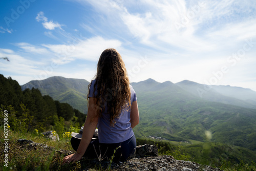 Billede på lærred Rear view of a female traveler sitting quietly on a mountain viewpoint surrounded by scenic summer nature