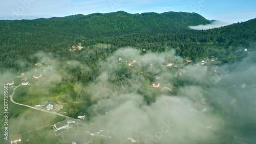 Cuadro en lienzo A scenic aerial view captures a tranquil valley surrounded by dense forest, with clouds gently rolling over the trees, offering a serene glimpse of nature's beauty