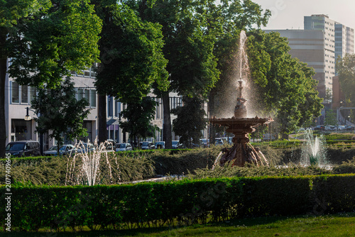 An ornate cast-iron fountain sprays water in a green city park with classic buildings in the background.