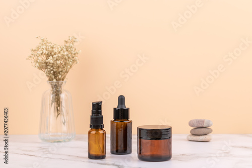 Horizontal front view with two amber glass bottles and cosmetic jar on marble table with zen stones and glass vase with dried flowers. Minimalist peach background with copy space