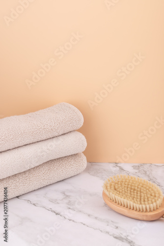 Vertical composition with stacked beige towels and wooden body brush on marble surface. Peach background with copy space. Concept of self care and natural lifestyle