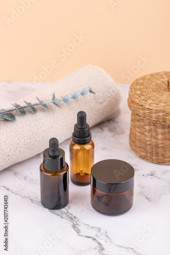 Amber glass cosmetic bottles with dropper and black lid beside rolled towel, eucalyptus branch and wicker basket. Peach background. Vertical minimal image with copy space