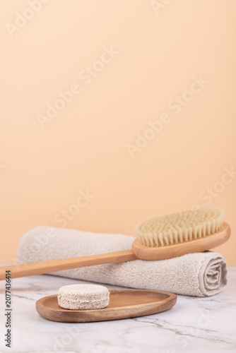 Front view vertical of wooden body brush, solid shampoo bar on tray and beige towel on marble base with peach background. Minimal spa style with copy space